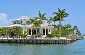 A luxury two-story waterfront estate with a metal roof and white columns, surrounded by tropical palm trees along a sea-walled canal with a docked yacht in Clearwater Beach, Florida.