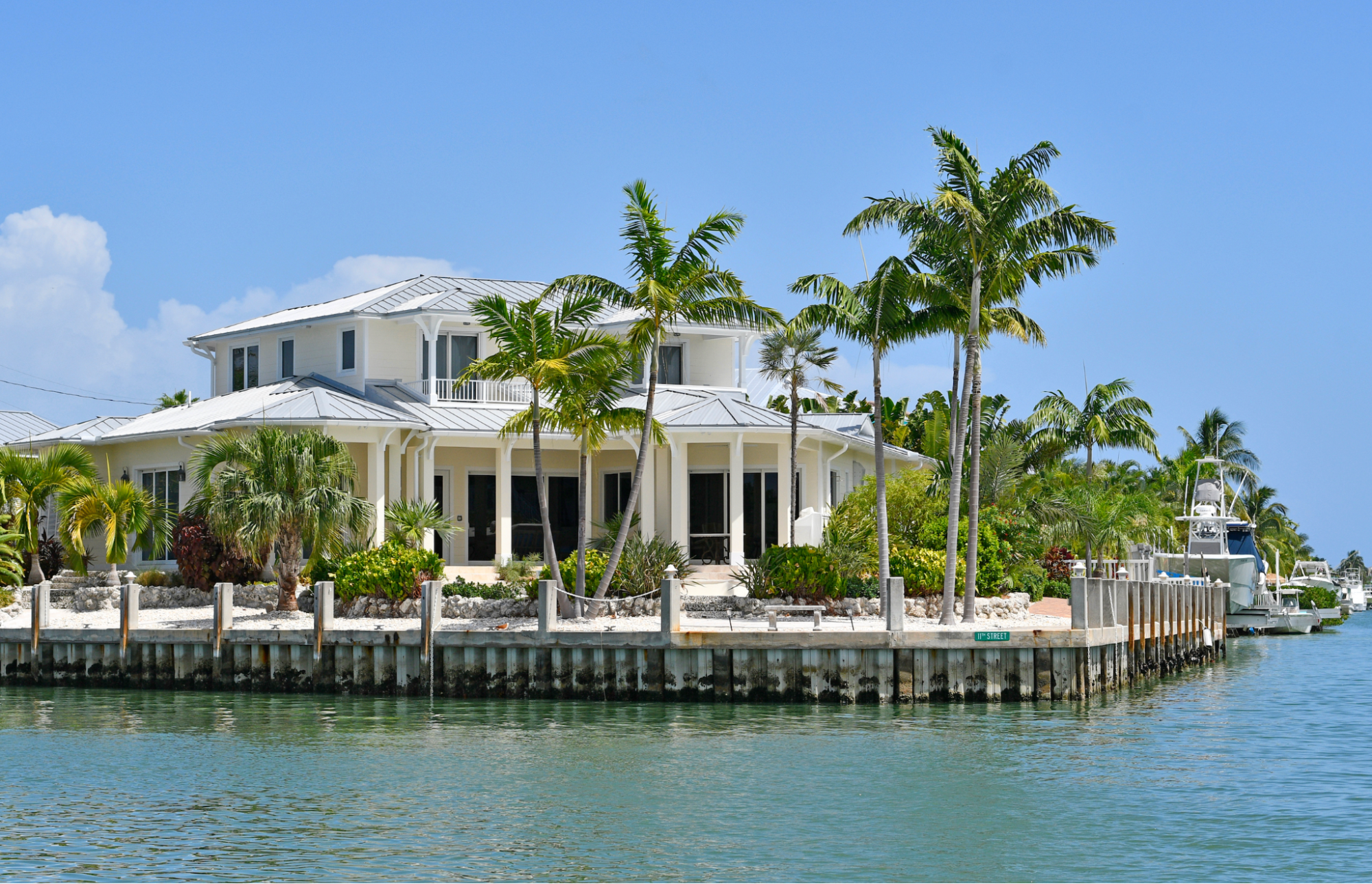 A luxury two-story waterfront estate with a metal roof and white columns, surrounded by tropical palm trees along a sea-walled canal with a docked yacht in Clearwater Beach, Florida.